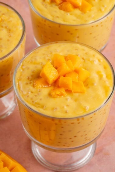 small glass cups of mango sago next to fresh mango on pink table