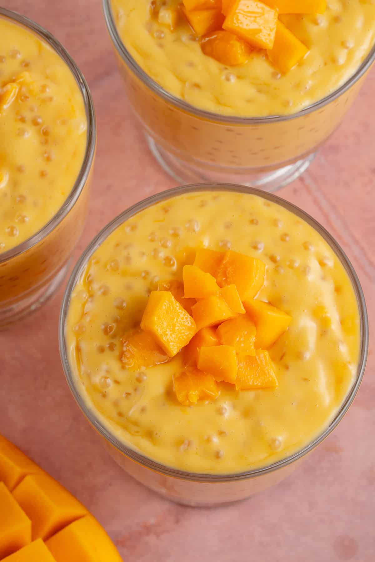 overhead photo of small glass cups of mango sago next to fresh mango on pink table