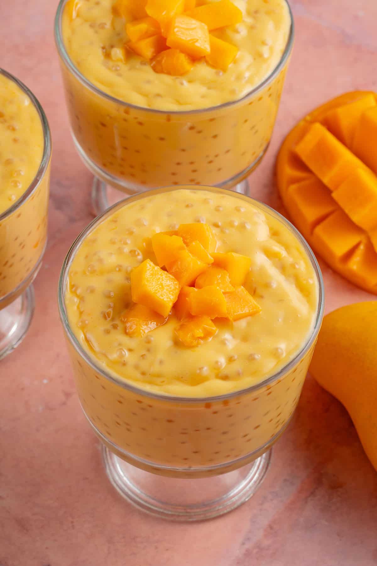 small glass bowls of mango sago next to fresh mango on pink table