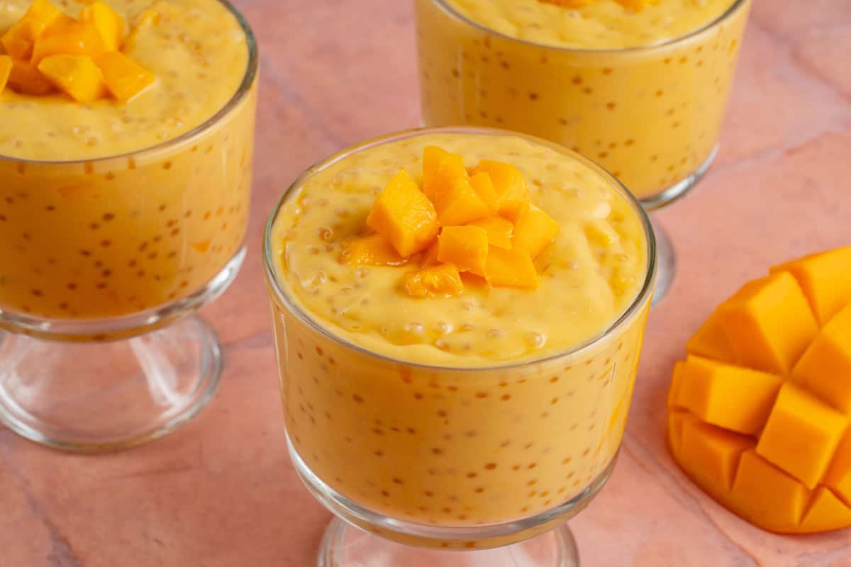 small glass bowls of mango sago next to fresh mango on pink table
