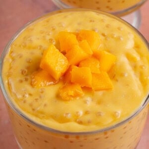 close up of glass cup of mango sago next to fresh mango on pink table