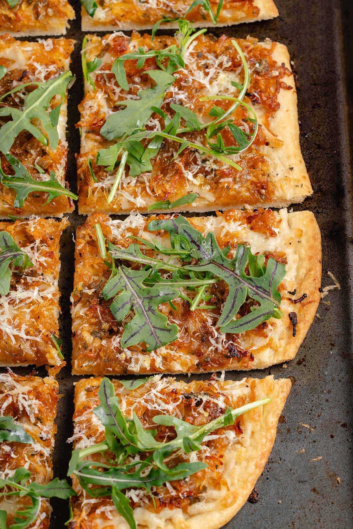 sliced vegan caramelized onion flatbread on dark baking sheet with arugula