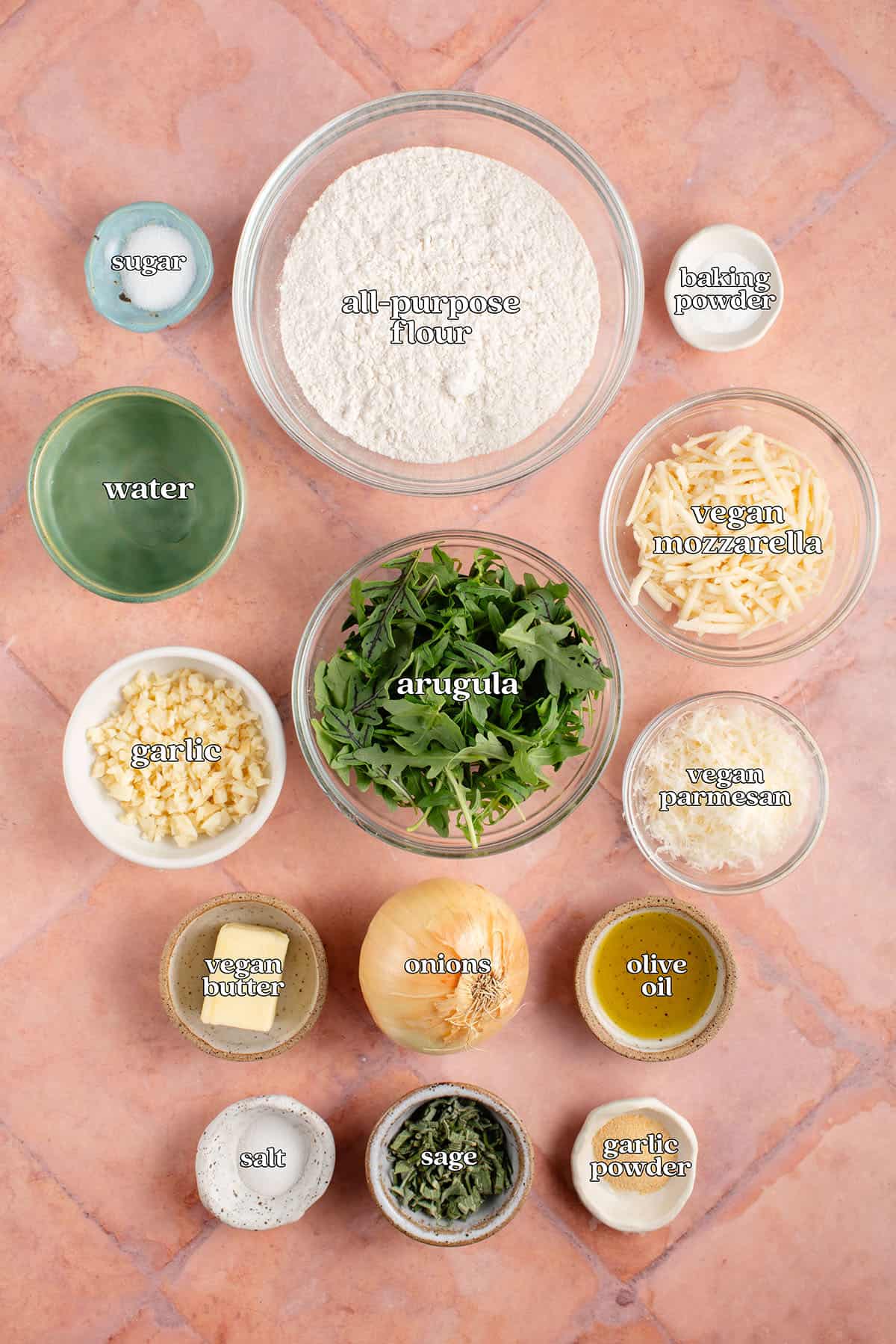 labelled ingredients for Easy Caramelized Onion Flatbread: sugar, all-purpose flour, baking powder, water, arugula, vegan mozzarella, garlic, vegan parmesan, vegan butter, onions, olive oil, salt, sage and garlic powder