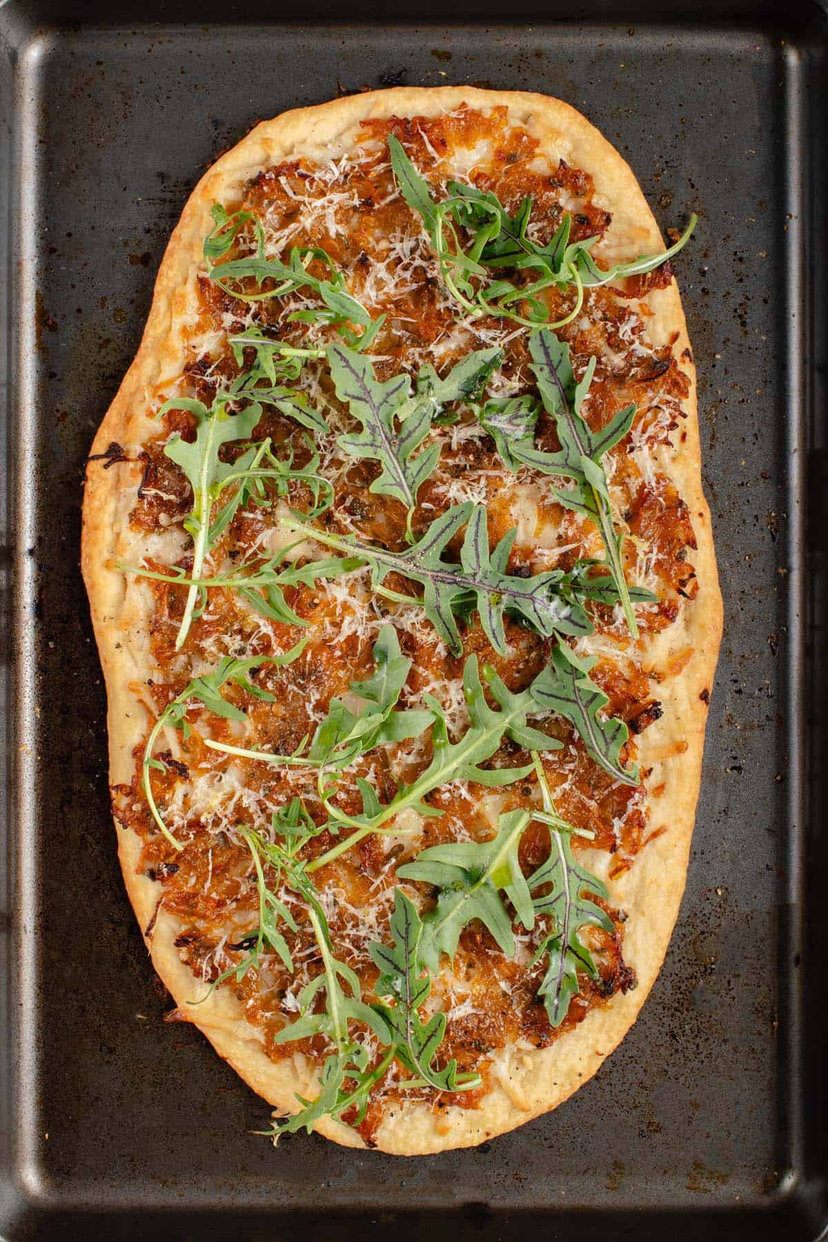 overhead photo of vegan caramelized onion flatbread on dark baking sheet with arugula