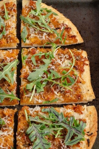 sliced vegan caramelized onion flatbread on dark baking sheet with arugula