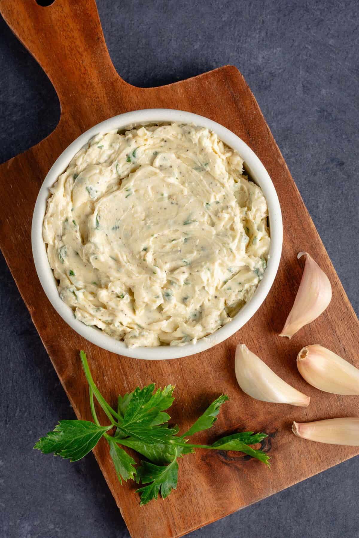bowl of garlic butter on wooden platter with cloves of garlic and parsley