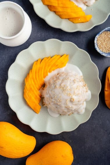 mound of mango sticky rice oatmeal topped with sauce and sesame seeds on a plate and a side of sliced mango
