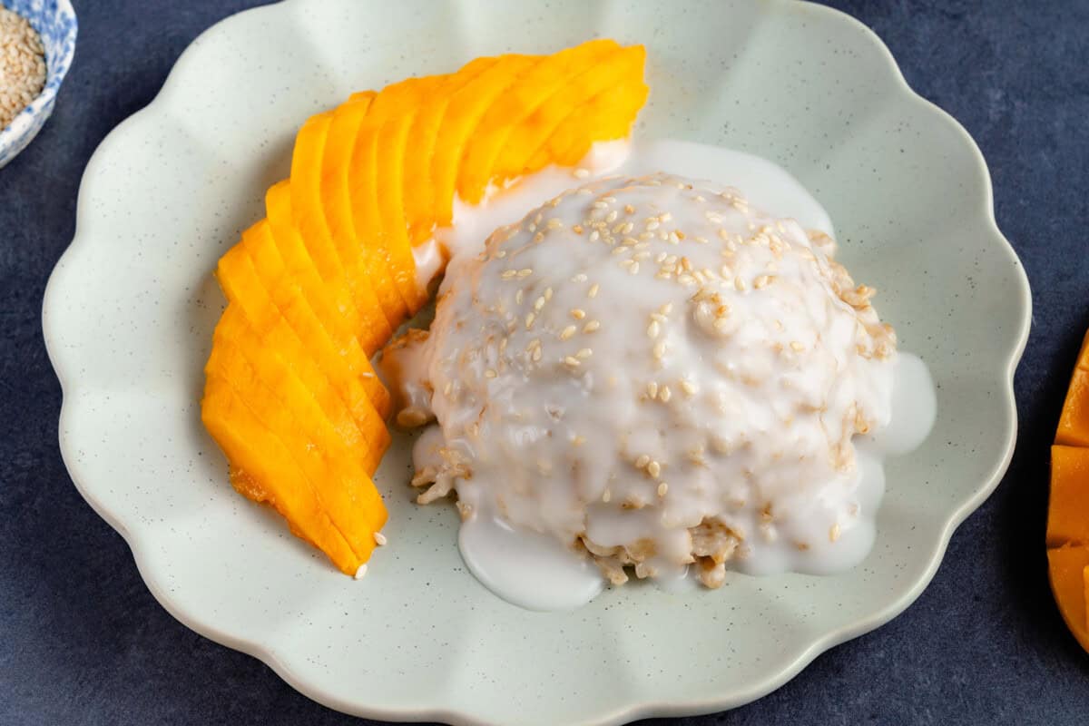 mound of mango sticky rice oatmeal topped with sauce and sesame seeds on a plate and a side of sliced mango