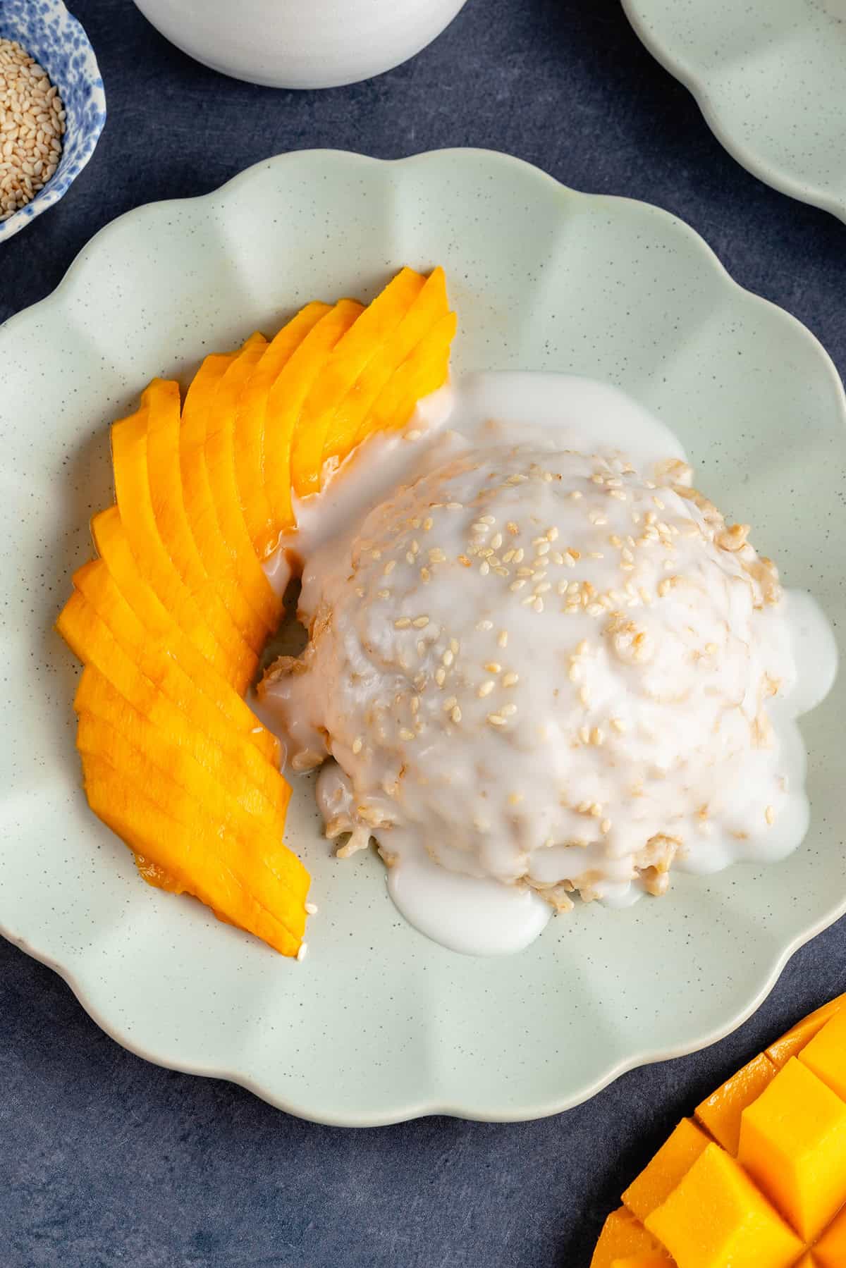 mound of mango sticky rice oatmeal topped with sauce and sesame seeds on a plate and a side of sliced mango