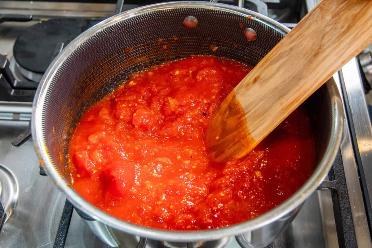 breaking whole peeled tomatoes with wooden spatula in pot