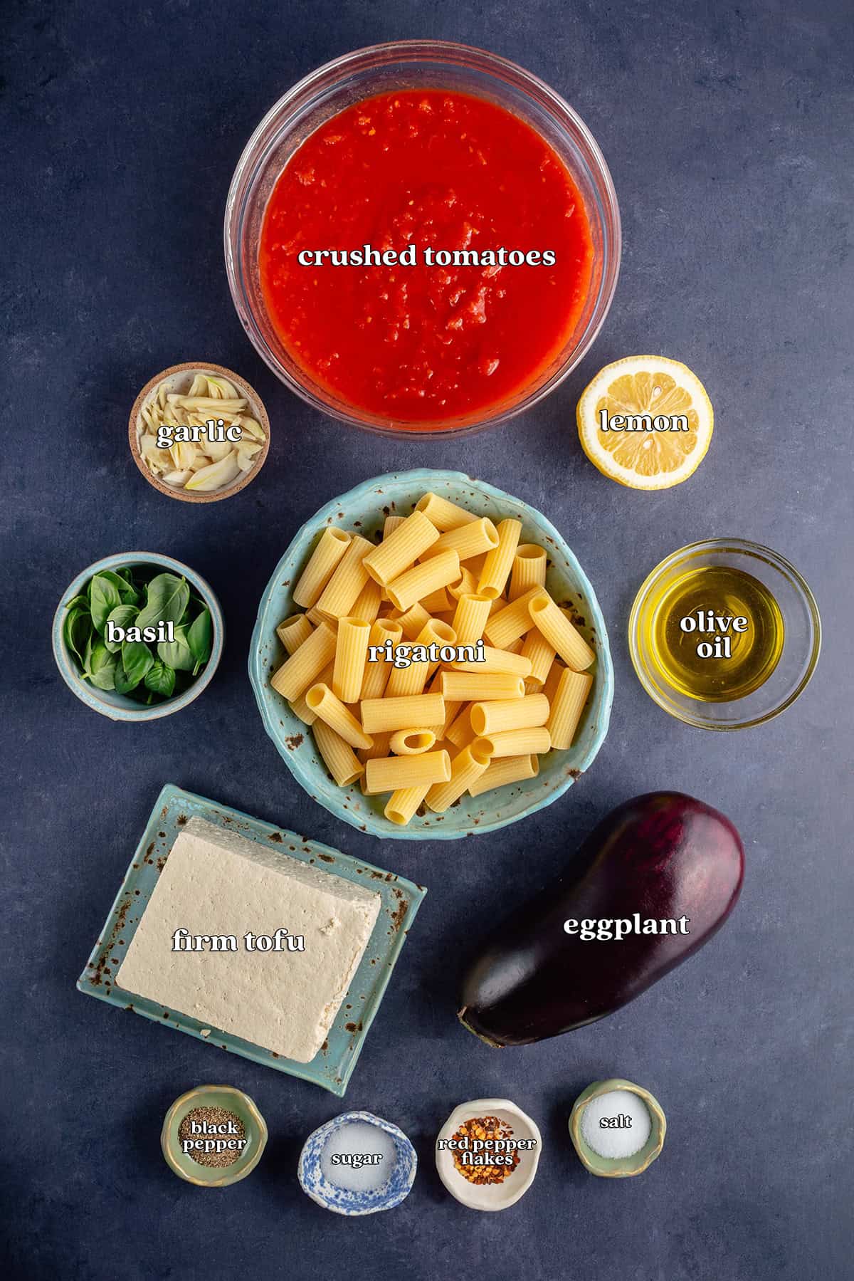 labeled ingredients for Pasta alla Norma (Eggplant Pasta): crushed tomatoes, garlic, lemon, basil, rigatoni, olive oil, firm tofu, eggplant, black pepper, sugar, red pepper flakes and salt