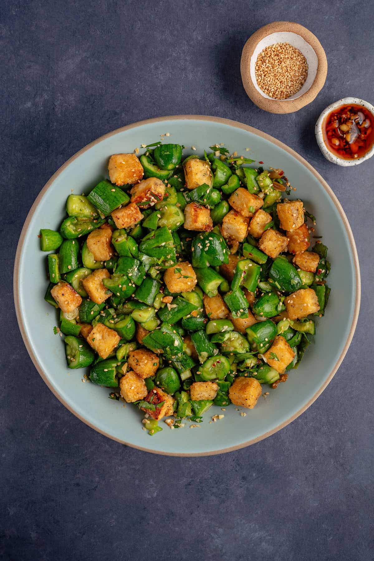 smashed cucumber salad in a big blue bowl with side of sesame seeds and chili crisp