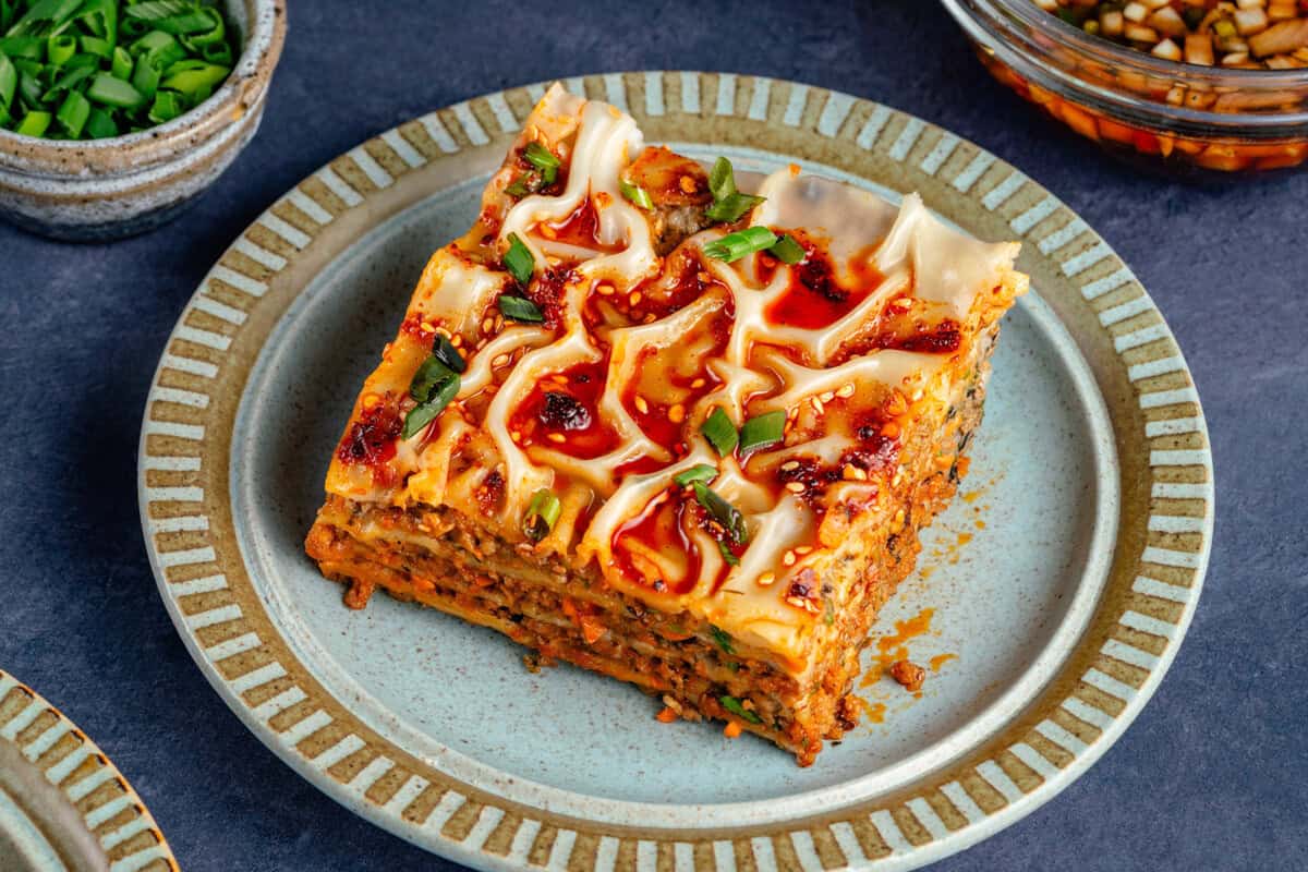 slice of lumpia-inspired dumpling lasagna on blue plate topped with chili oil, sawsawan, and green onions