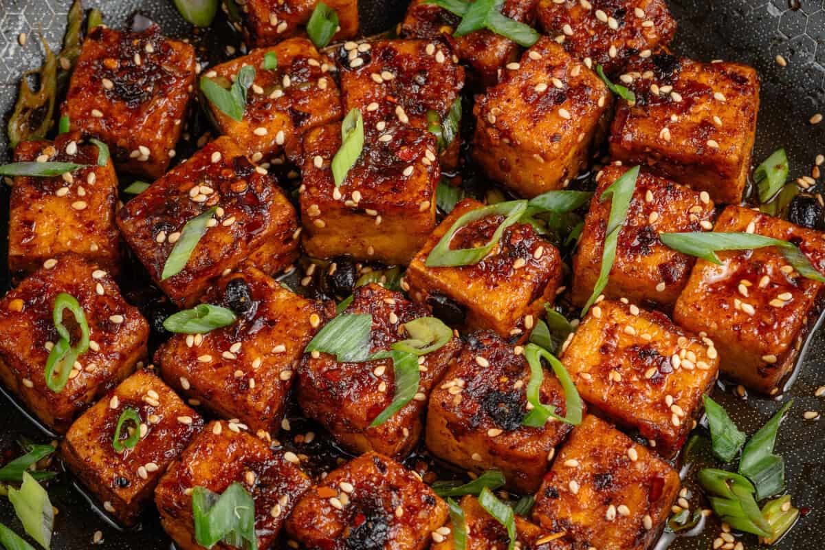 horizontal photo of Chili Crisp Tofu cubes in pan with sliced green onions and toasted sesame seeds