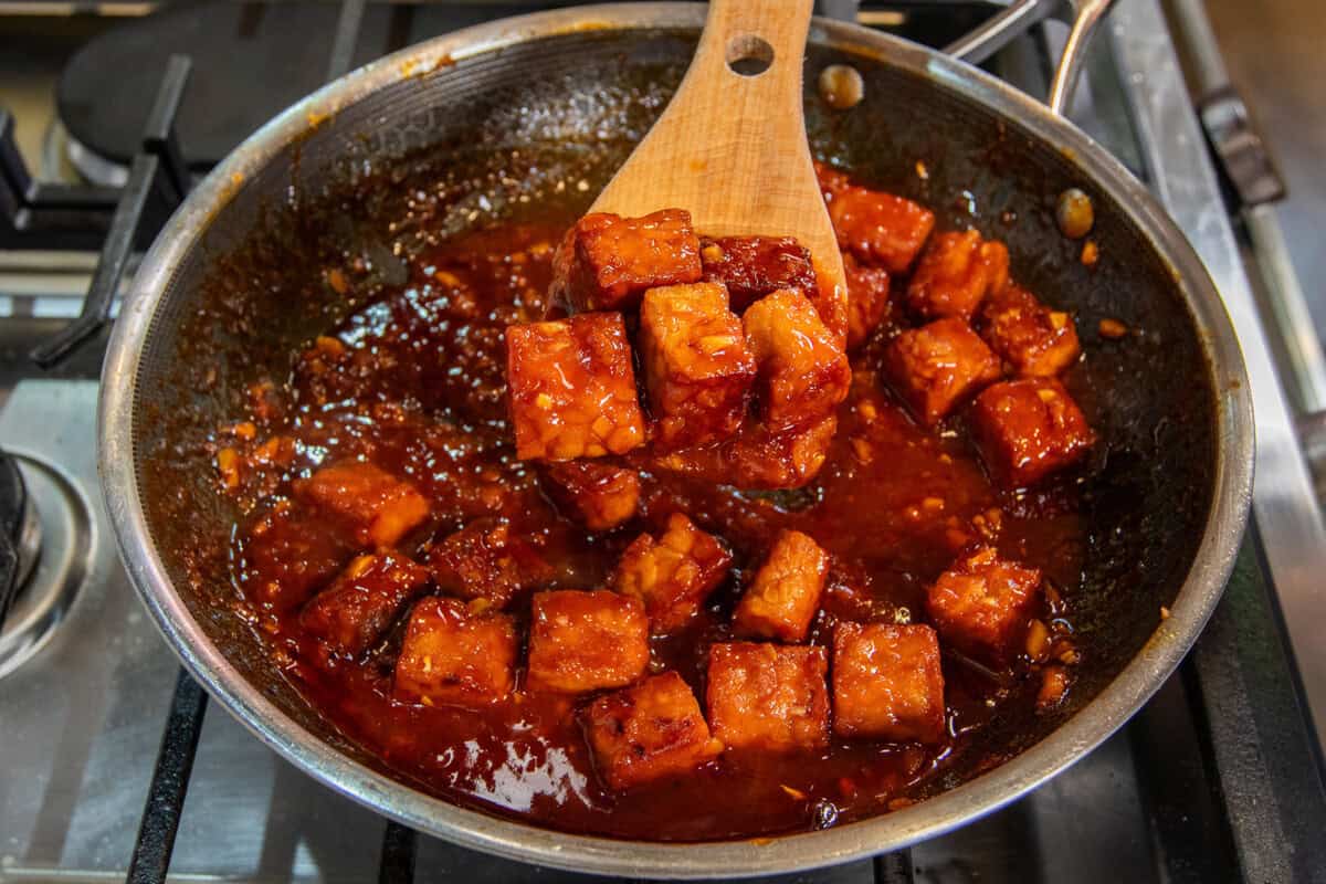 scooping gochujang braised tempeh out of a pan with a wooden spoon