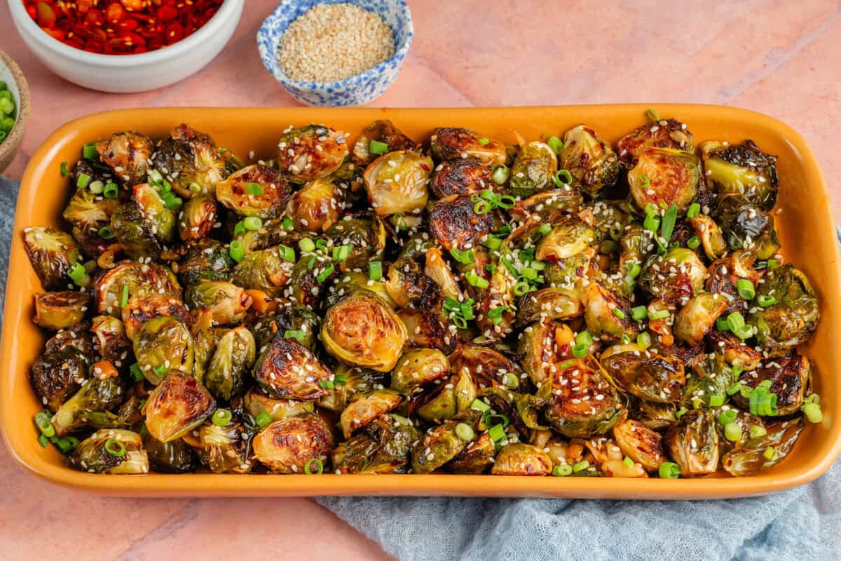 maple chili crisp brussels sprouts in yellow platter and chili crips, green onions, and sesame seeds on the side