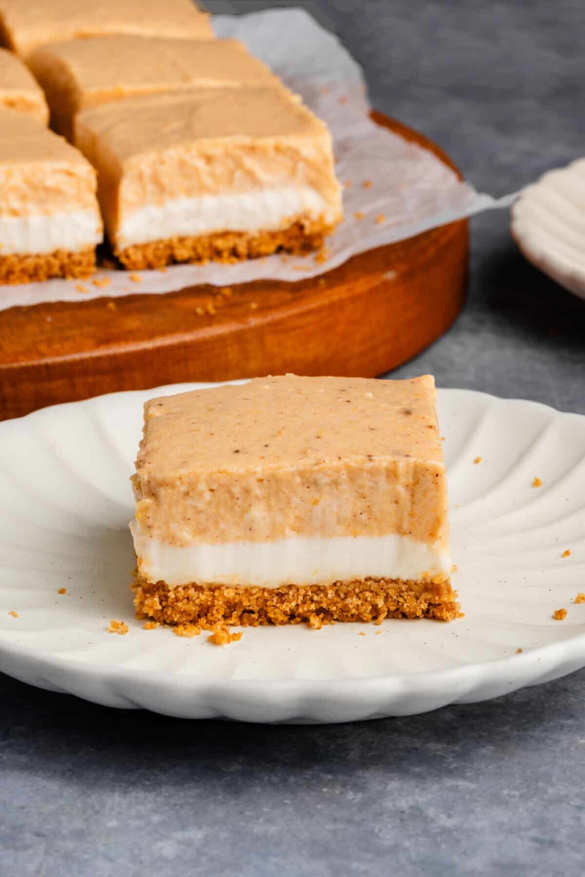 vertical photo of a Pumpkin Cheesecake Bar on a white scalloped plate with additional bars on a platter in the background
