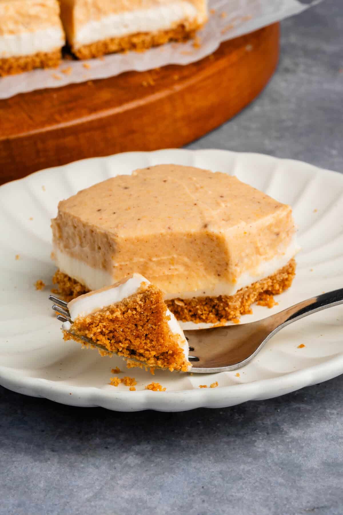 a cut Pumpkin Cheesecake Bar with a fork on a white scalloped plate with additional bars in the background