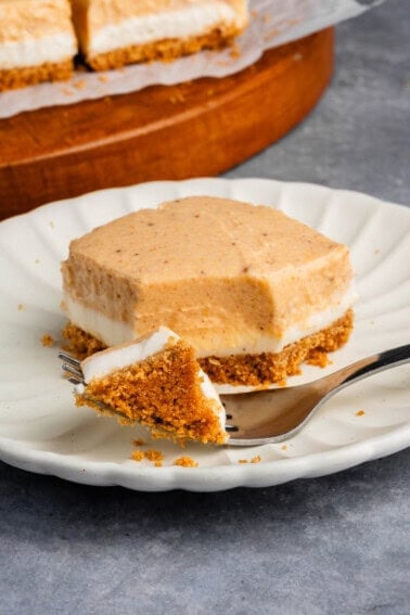 a cut Pumpkin Cheesecake Bar with a fork on a white scalloped plate with additional bars in the background