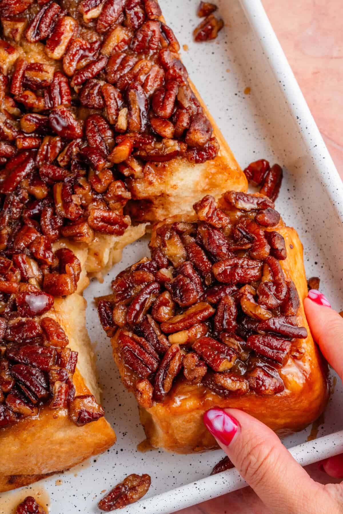 maple pecan sticky buns in a sheet pan with one broken off being held by a hand